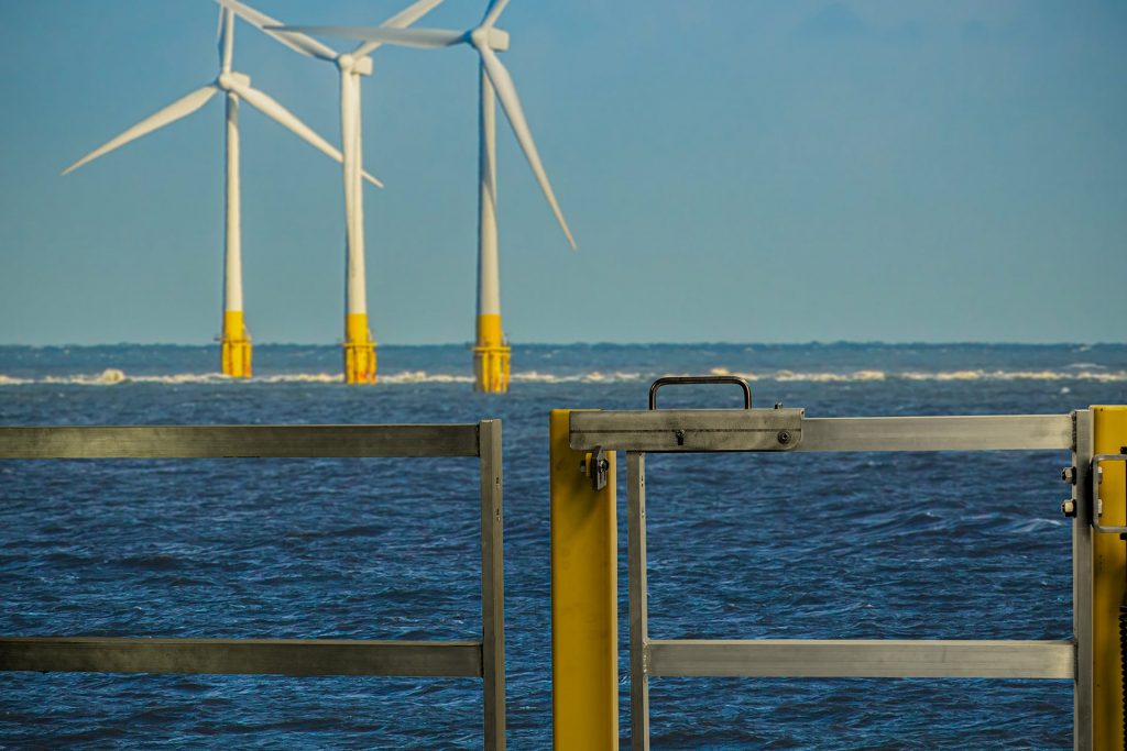 Single gate latch  hardware for offshore in yellow gate with three wind turbines in the sea in the background.